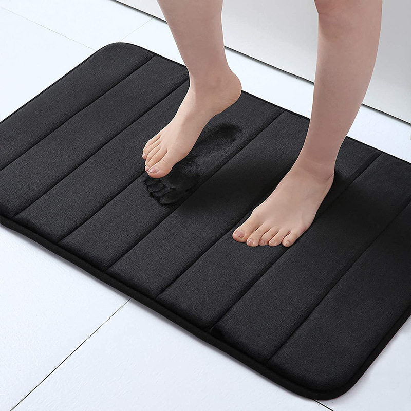 Black bath mat with feet stepping onto it on a white floor.