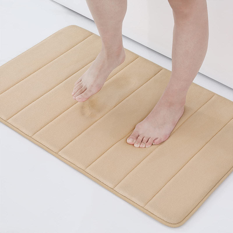 A beige rectangular bath mat with vertical channel stitching, shown with a person's bare feet stepping onto it in a clean bathroom setting.