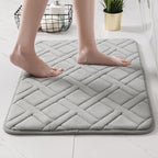 A light gray quilted bath mat with a diamond pattern, shown with bare feet standing on it in a bright bathroom setting.