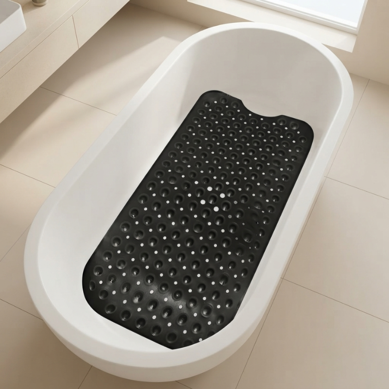 A black textured bath mat with raised circular bumps, placed inside a white freestanding bathtub on a light beige tiled floor.