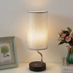 Table lamp with a textured shade on a white surface, next to a framed picture and a vase with flowers.