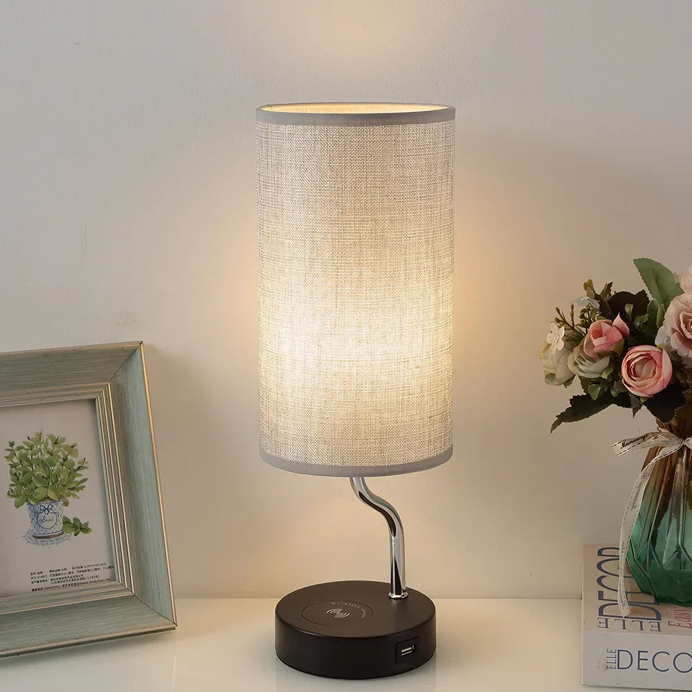 Table lamp with a textured shade on a white surface, next to a framed picture and a vase with flowers.