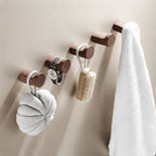Four dark brown wooden wall hooks mounted on a beige wall, holding a white bath sponge, a watch, a brush, and a white towel.