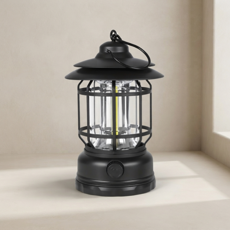 A black lantern-style LED camping light with a protective cage and hanging loop, shown on a light beige surface against a neutral beige background.