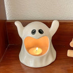 A ceramic ghost-shaped candle holder with a small tea light candle inside, displayed on a wooden surface against a textured wall.