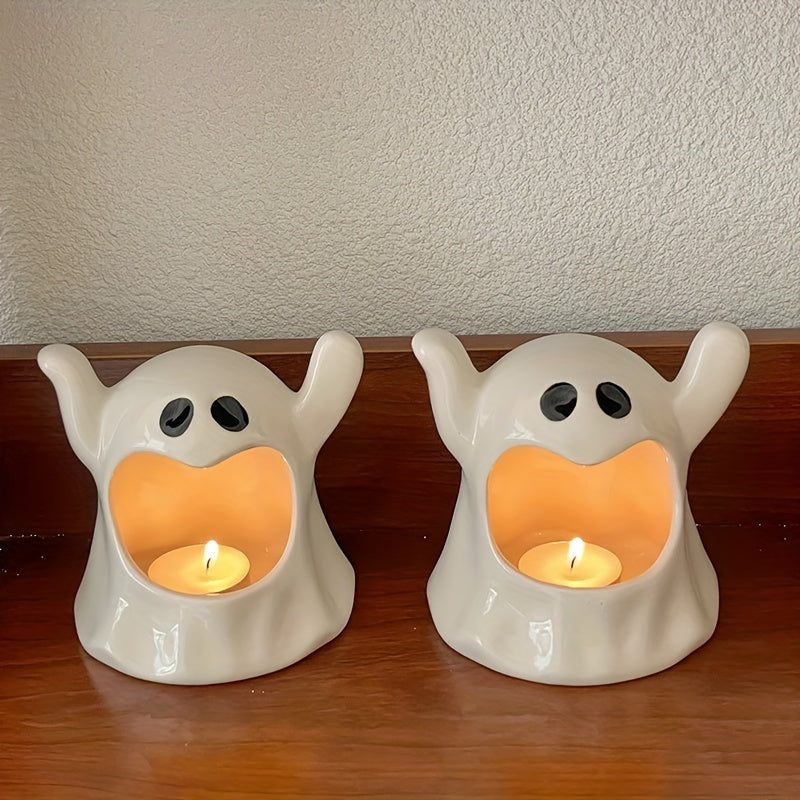 Two cute ghost-shaped ceramic tea light candle holders with raised hands, displayed side-by-side on a wooden surface against a textured beige wall. Each ghost features a black face with eyes and holds a glowing tea light candle inside its open mouth.