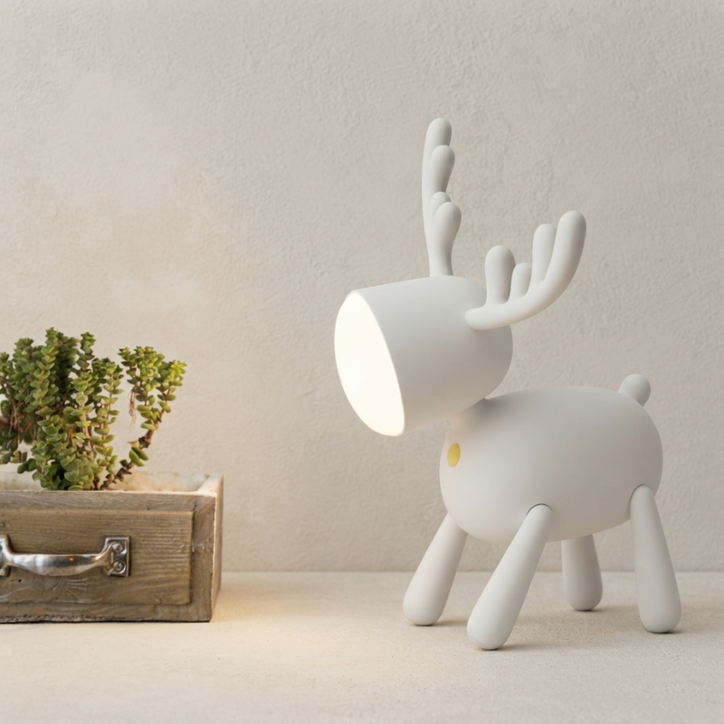 A white reindeer-shaped table lamp with a cylindrical head and small yellow accent, shown on a tabletop against a textured beige wall next to a wooden planter.
