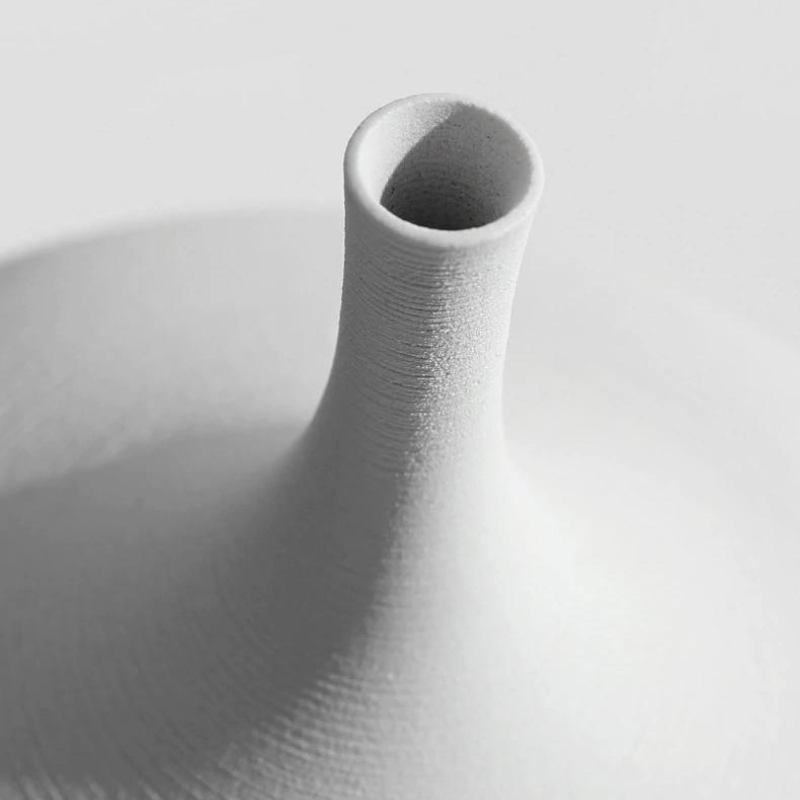 A close-up studio shot of a matte white ceramic vase with a flared base and a narrow cylindrical neck, showing fine horizontal texture and a soft shadow on a light gray background.