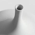 A close-up studio shot of a matte white ceramic vase with a flared base and a narrow cylindrical neck, showing fine horizontal texture and a soft shadow on a light gray background.