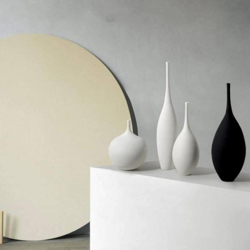 Four minimalist matte ceramic vases in white and black, arranged on a white pedestal against a gray wall with a large cream-colored circular backdrop.