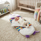 A baby wearing a cream bodysuit sleeps on a plush white unicorn-shaped baby blanket with purple ears and a rainbow-striped border, placed on a beige shag rug in a nursery.