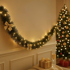 A festive Christmas garland with warm white lights and gold bow accents draped across a cream wall, with a decorated Christmas tree and wrapped gift in the background.