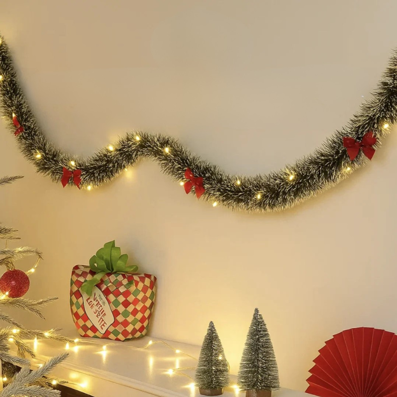 A festive Christmas garland with silver tinsel, warm white string lights, and red bows is draped across a beige wall above a mantel decorated with a wrapped gift, miniature trees, and warm lights.