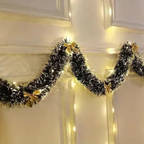 Black tinsel Christmas garland with warm white string lights and small gold bows, draped across a cream-colored paneled wall.