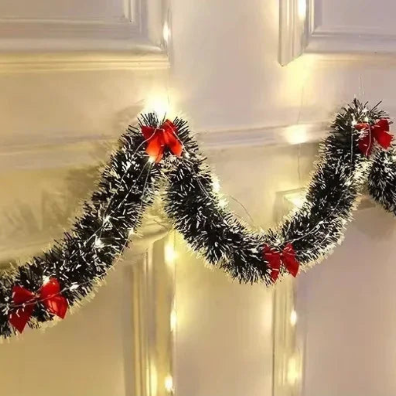 Black tinsel Christmas garland with white faux snow and red ribbon bows, draped across a cream-colored paneled door with warm string lights.