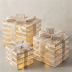 Three stacked gift box lanterns in different sizes, decorated with beige burlap ribbons and bows, glowing with warm internal fairy lights against a neutral beige background.