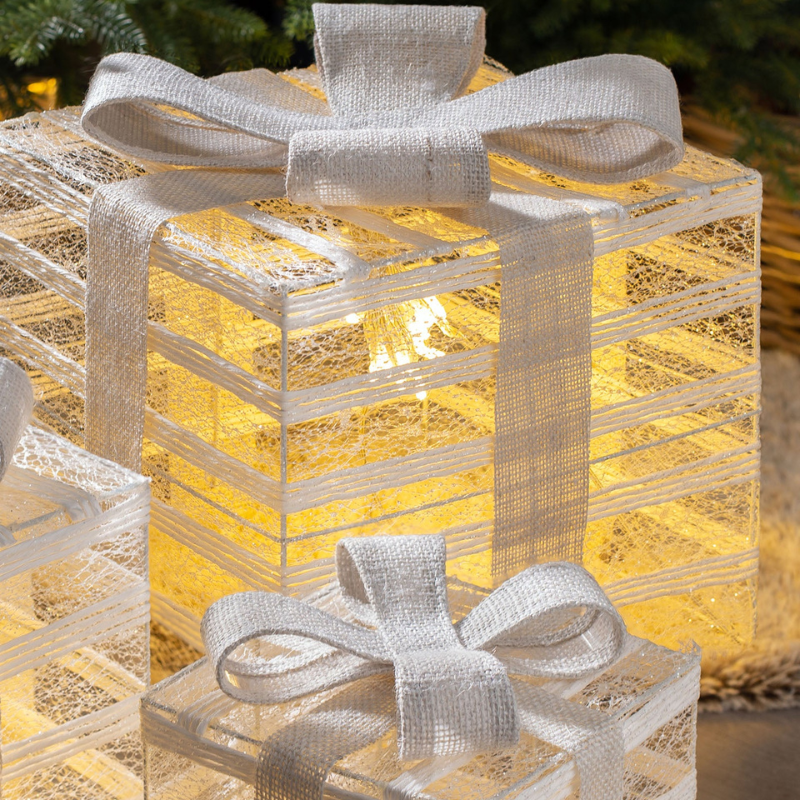 A decorative gift box lantern with a glowing warm yellow light inside, wrapped in a sheer white burlap ribbon and bow, shown on a softly blurred outdoor background.