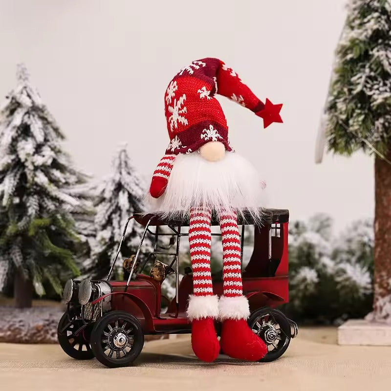 A festive Christmas gnome decoration with a red and white striped knit hat, white beard, and striped legs, sitting on top of a small red vintage car against a snowy tree backdrop.
