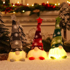 Three Christmas gnome floor lights arranged in a row on a snowy surface, glowing warmly. The gnomes have tall knit hats in gray, red, and green with white snowflakes, white beards, and small gray, red, and green feet.