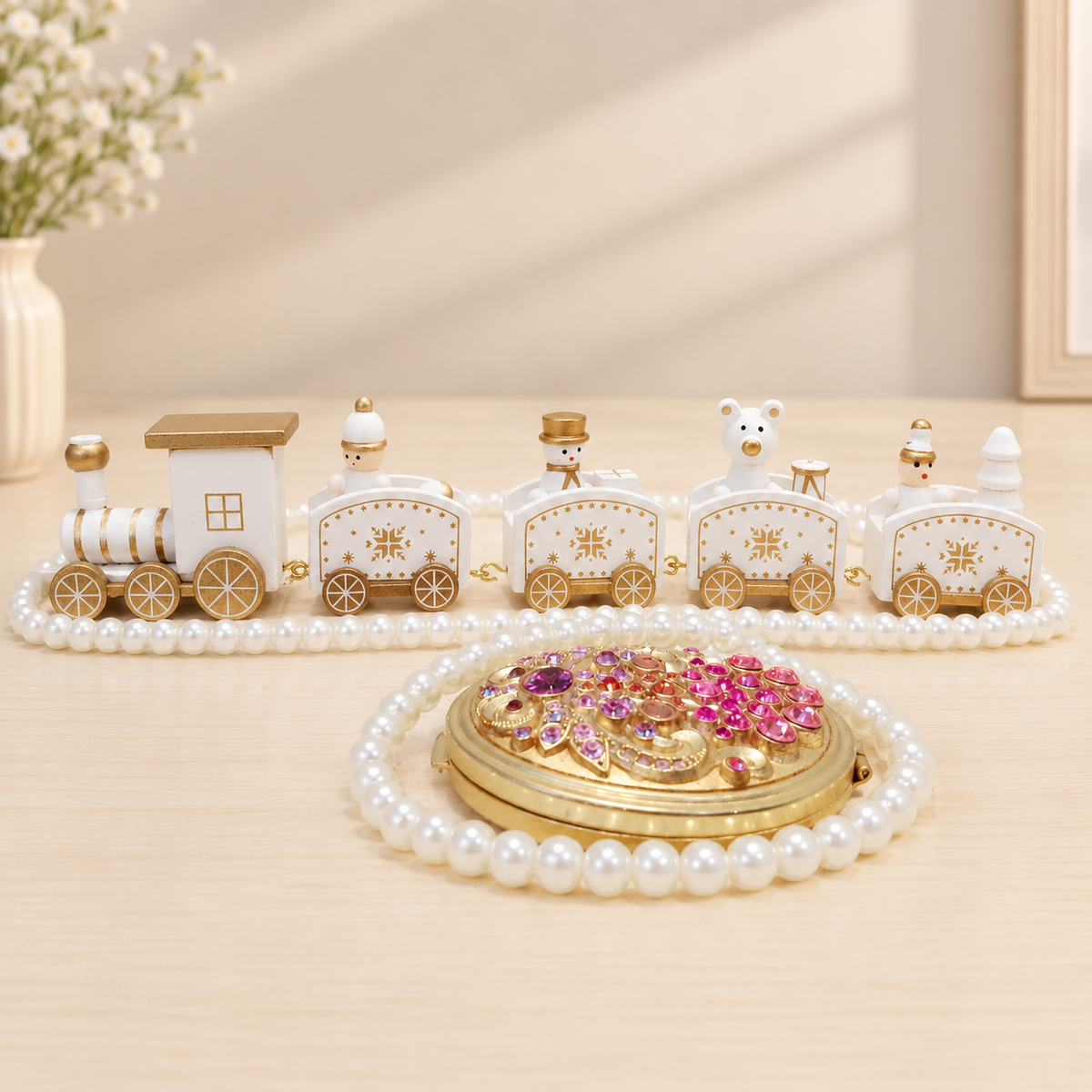 A decorative jewelry organizer set featuring a small white and gold train made of faux pearls and gold-toned metal, paired with a round gold compact mirror embellished with pink and purple rhinestones, all arranged on a light beige surface.