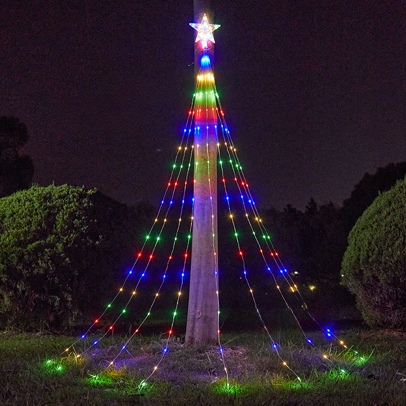 A tall outdoor Christmas tree light display on a pole at night, with a glowing star on top and multicolored string lights forming a tree shape on the pole and spreading out onto the grass.