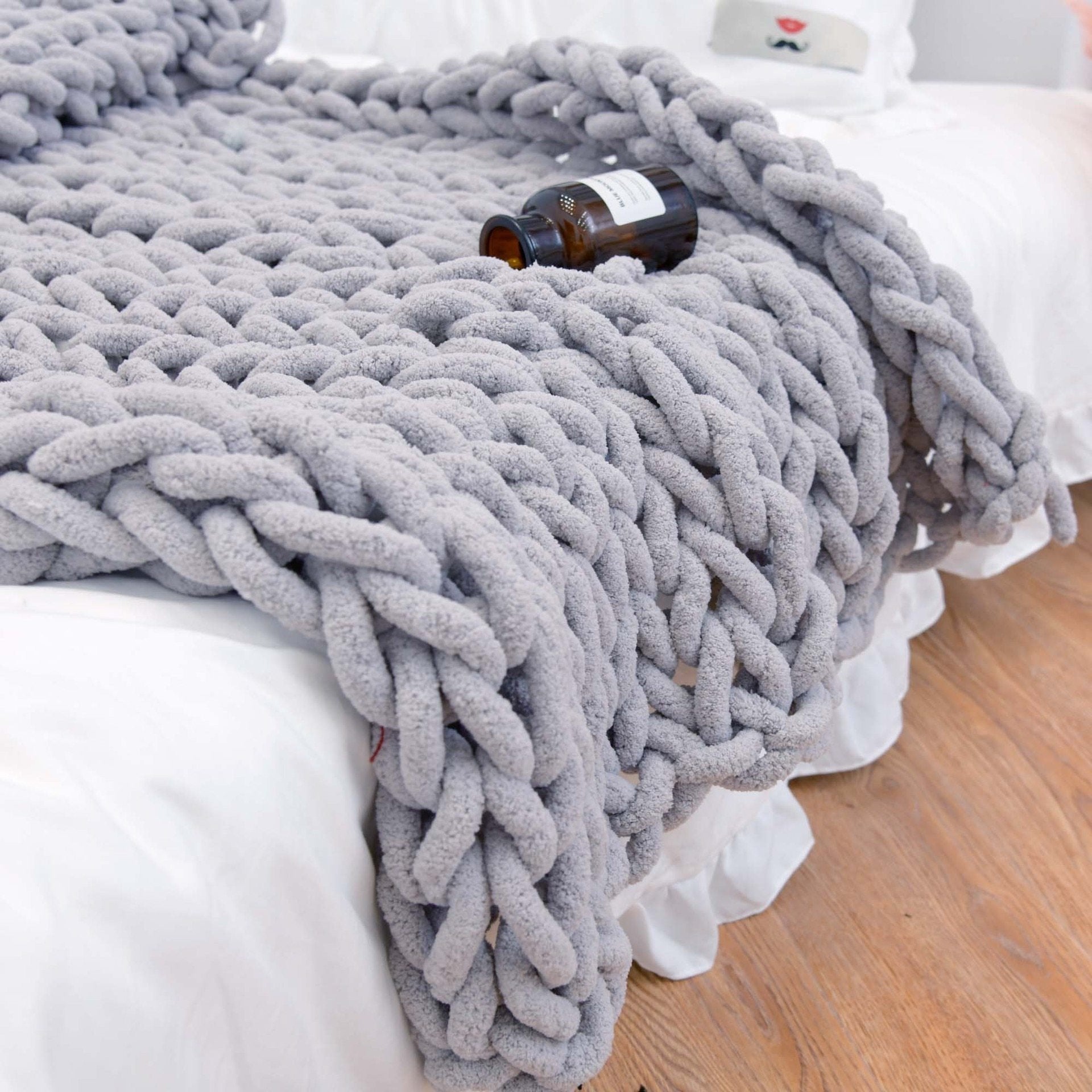 Gray chunky knit blanket draped over a bed with a small bottle on top.