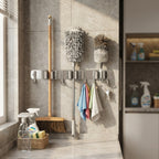 A wall-mounted metal broom and duster holder in a tiled bathroom or utility space, holding a broom, two dusters, and hanging towels. Cleaning supplies and a basket sit on the counter nearby.