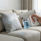A handheld white misting device with a clear bottle of blue liquid is being used to spray a light gray sofa cushion in a living room setting.
