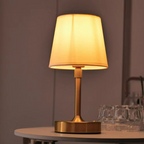 Gold table lamp with a beige shade on a round table against a purple wall.