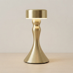 A modern table lamp with a brushed brass/gold metallic finish, featuring a flared base and a cylindrical shade that glows with warm light, set against a neutral beige background.