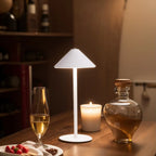 A modern white table lamp with a conical shade and circular base stands on a wooden table, lit to cast a warm glow. Surrounding it are a lit candle, a glass of white wine, and a clear glass decanter with amber liquid, set against a softly blurred indoor background.
