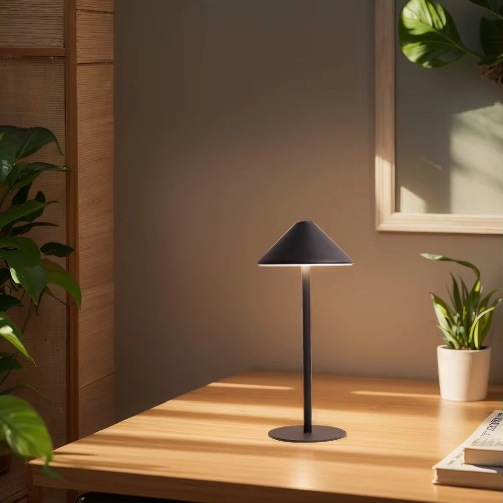 A modern black table lamp with a conical shade and round base sits on a wooden desk in a warm, sunlit room with plants and a framed mirror in the background.