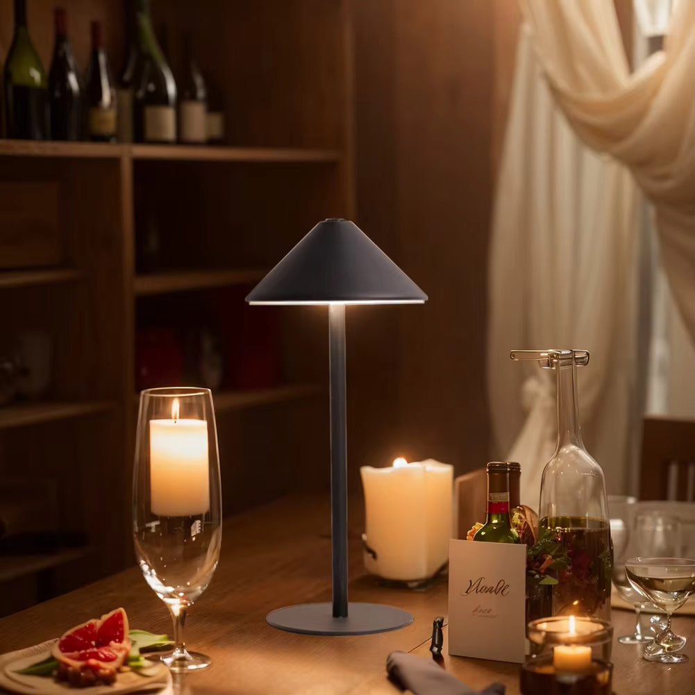 A modern table lamp with a dark gray conical shade and circular base, illuminated on a wooden dining table surrounded by candles, wine glasses, and bottles in a warmly lit room.