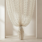 A cream-colored sheer curtain with small black embroidered dots, gathered and tied at the bottom in a window frame.