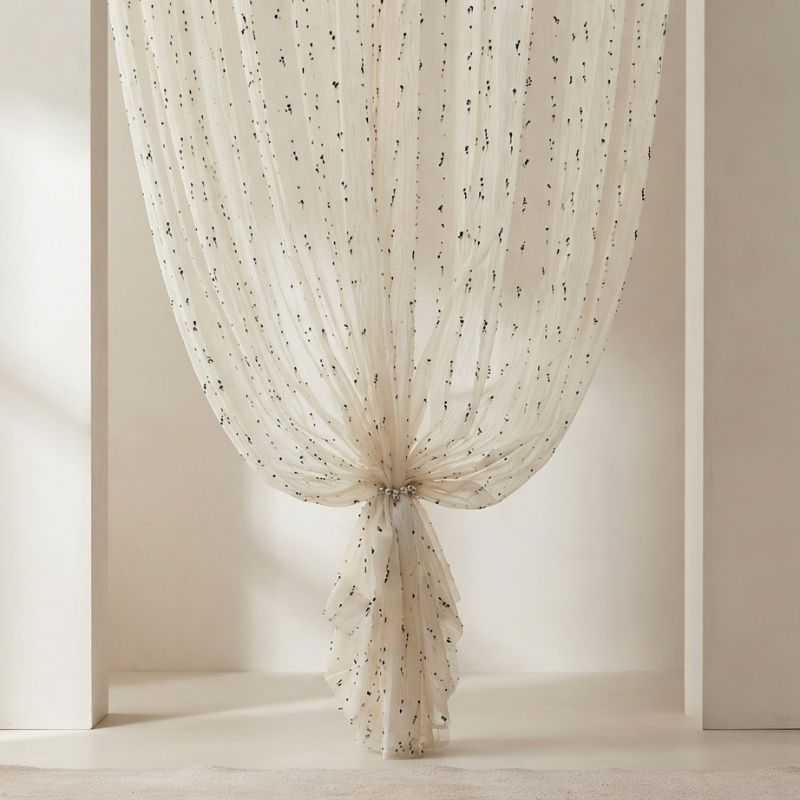A cream-colored sheer curtain with small black embroidered dots, gathered and tied at the bottom in a window frame.
