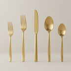 Five-piece gold flatware set with matte and satin finishes, including a dinner fork, salad fork, dinner knife, soup spoon, and teaspoon, standing upright on a light beige surface against a neutral background.