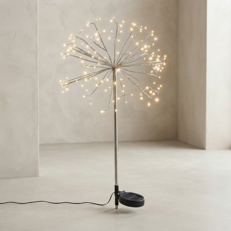 A dandelion-style floor lamp with glowing warm white fairy lights on thin metallic branches, mounted on a slim pole with a round black base and visible power cord, photographed in a minimalist beige room corner.