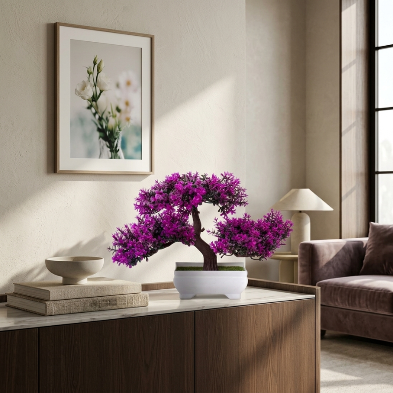 A potted bonsai-style plant with dense vivid purple flowers sits on a white marble-topped console in a styled living room. The room has beige walls, a framed floral print, a lamp, books, a bowl, and a brown upholstered chair by a window.