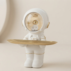 A white astronaut figurine holding a gold-colored tray, set against a soft beige background.