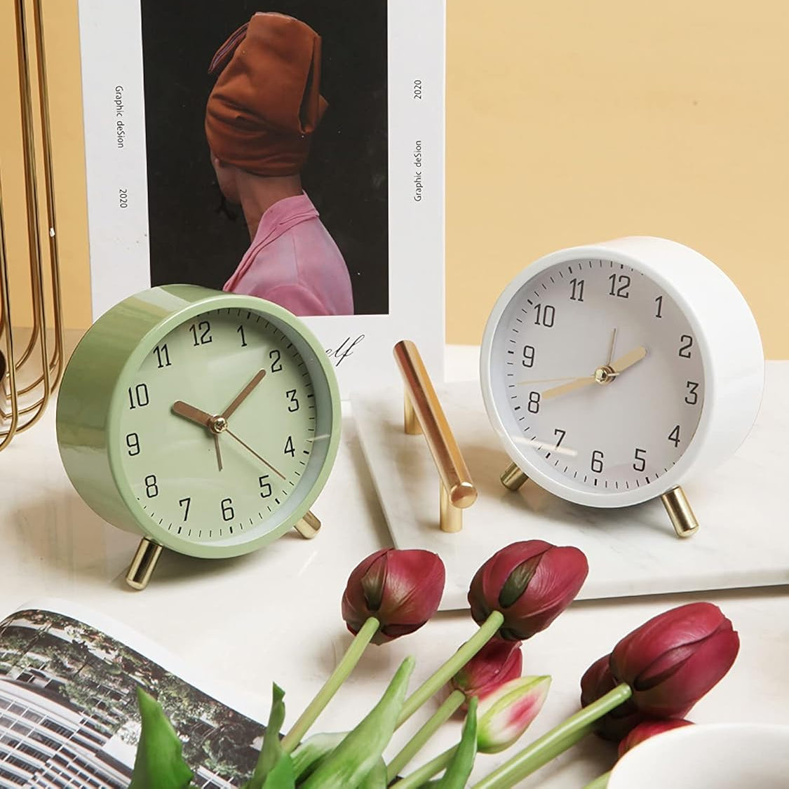Two alarm clocks, one green and one white, on a surface with tulips and a magazine.
