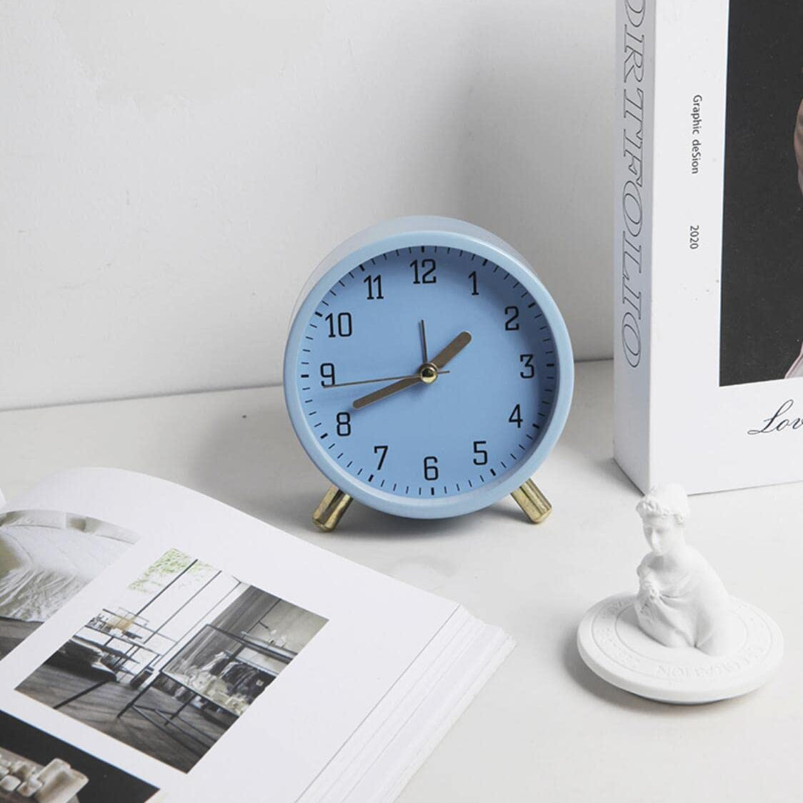 Light blue alarm clock on a white surface with a book and decorative item in the background