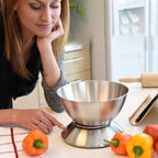 A woman in a kitchen looks down at a stainless steel countertop scale with a mixing bowl on top, surrounded by colorful bell peppers.