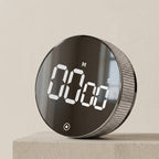 A round digital alarm clock with a glossy black face and a textured silver side, sitting on a light beige stone block against a neutral beige wall.