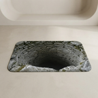 A rectangular bath mat with rounded corners featuring a printed design of a stone well opening into darkness, shown on a beige bathroom floor near a white tub.
