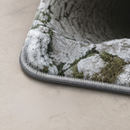 Close-up of a textured rug corner with a bark and moss pattern in gray, white, and green, shown on a beige floor.