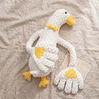 A white textured plush goose toy with yellow beak, feet, and wing details, posed on a beige linen fabric background next to a matching beige cloth.
