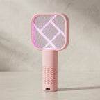 A pink handheld electric mosquito killer lamp with a square grid design, standing on a light gray surface against a textured light gray wall. The device is illuminated with a pink glow, and a USB charging port is visible on the bottom.