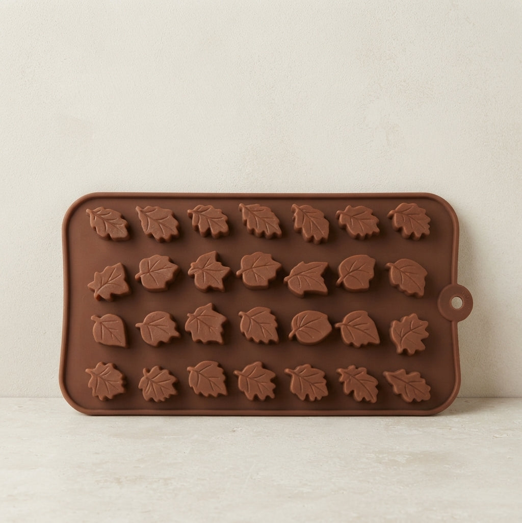 A rectangular brown silicone mold with embossed leaf-shaped cavities, shown upright against a light beige textured wall and surface.