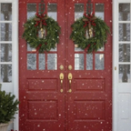 A pair of red double front doors with gold hardware, decorated with two green Christmas wreaths featuring red berries and gold bows, with snow gently falling in front.