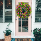 A vibrant, multicolored artificial flower wreath hangs on the pink front door of a white house, flanked by a potted green plant on the left and a flowering shrub on the right.